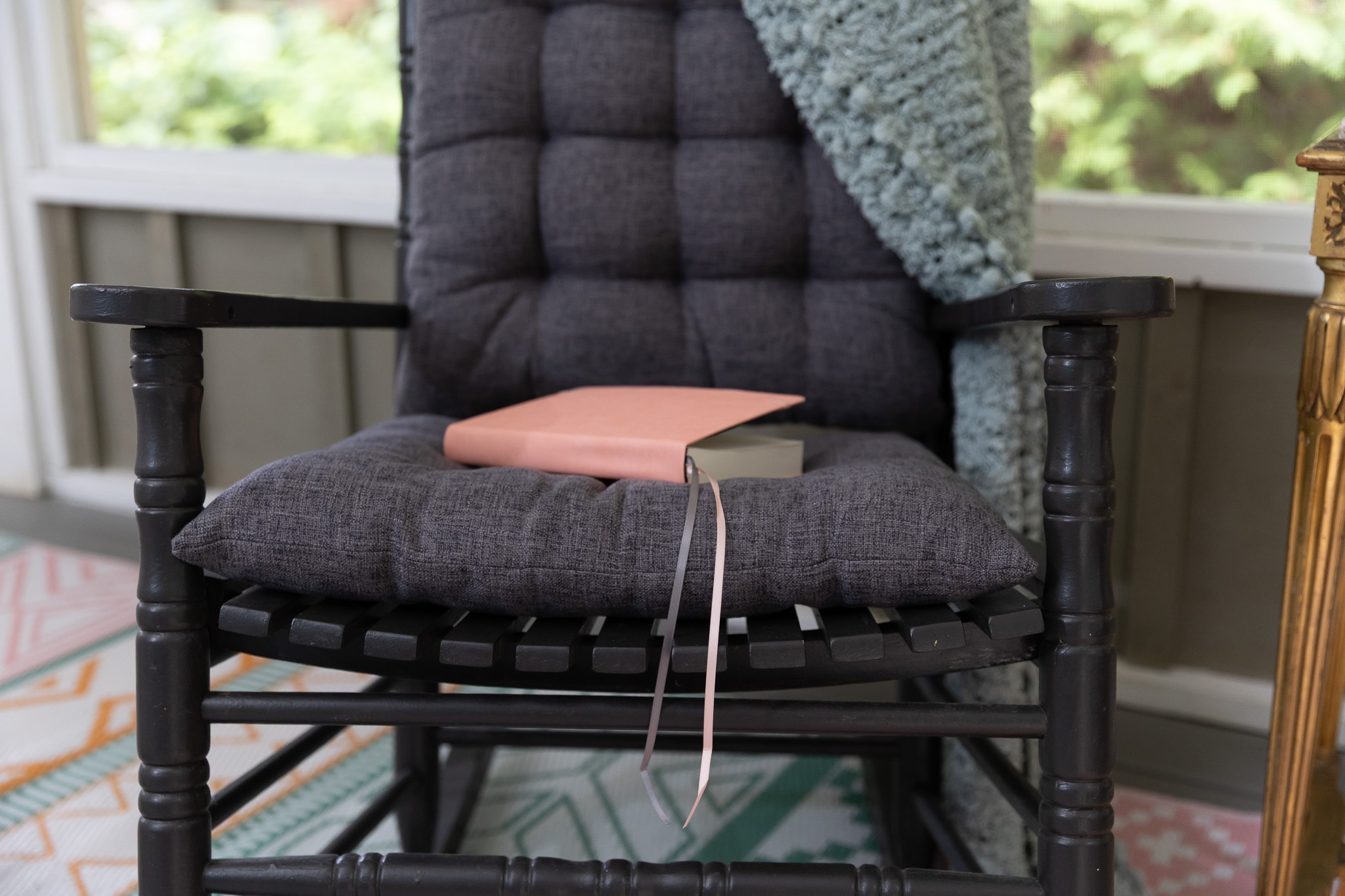 A black rocking chair with a teal throw blanket and a pink journal on the seat, on a sunlit porch